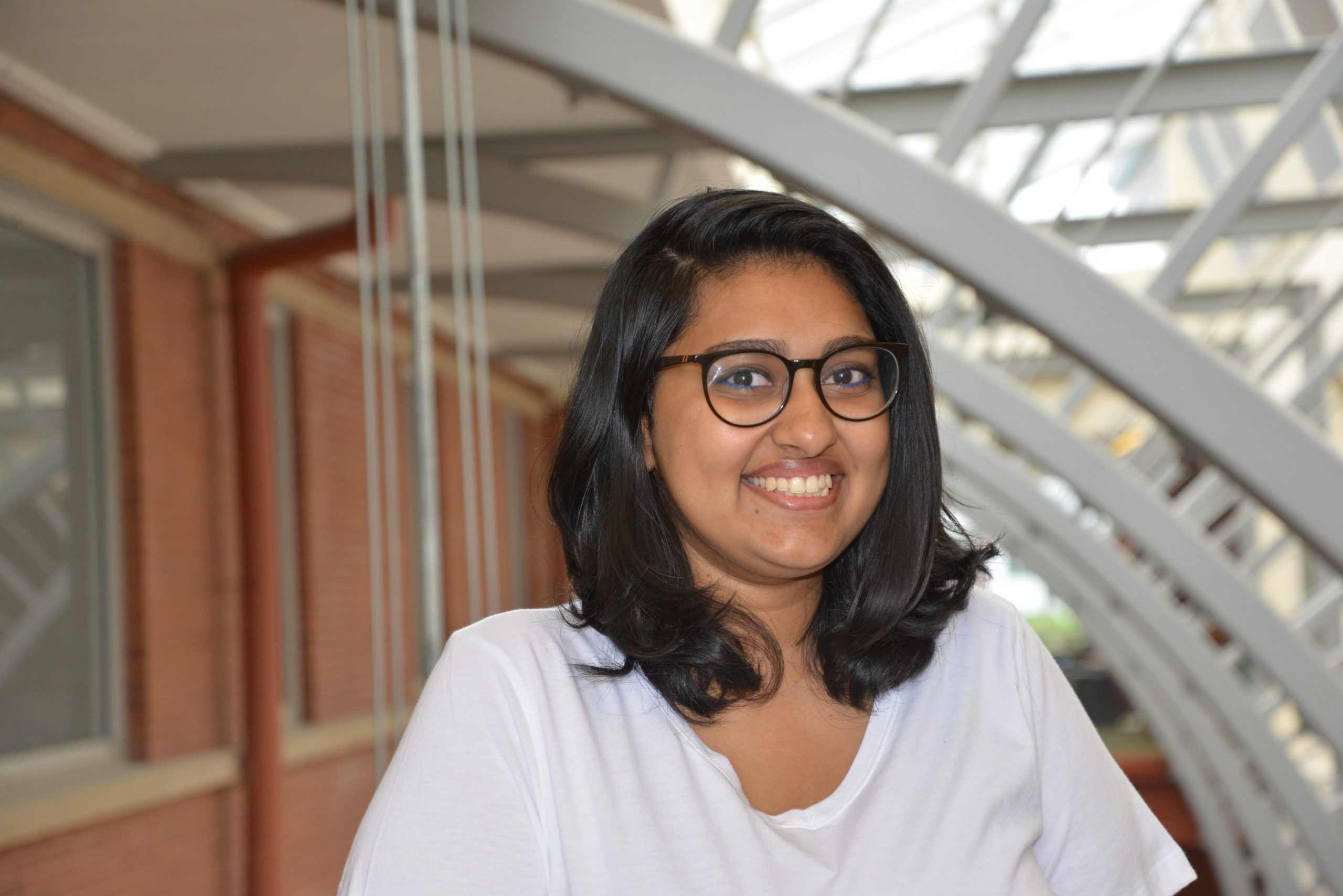 Vaishnavi Sivaprasad (a South Asian woman from India with dark brown shoulder-length hair and glasses) wearing a white t shirt, smiling into the camera