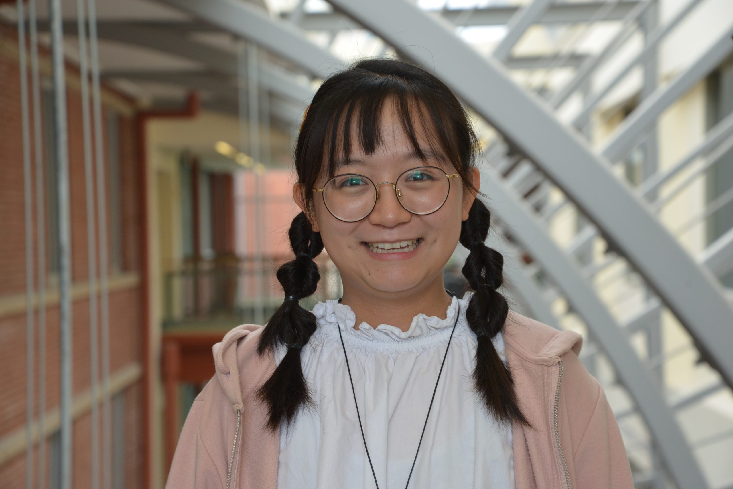 Yu-Shuang Liu (an East-Asian woman from Taiwan, with dark brown hair in pigtails, wearing glasses, a white shirt and a light pink hooded sweatshirt) smiling into the camera