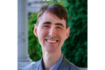 Noah Glaser Ph.D, (a white man with short dark brown hair and brown eyes, wearing a slate blue shirt and grey blazer), smiles into the camera side on